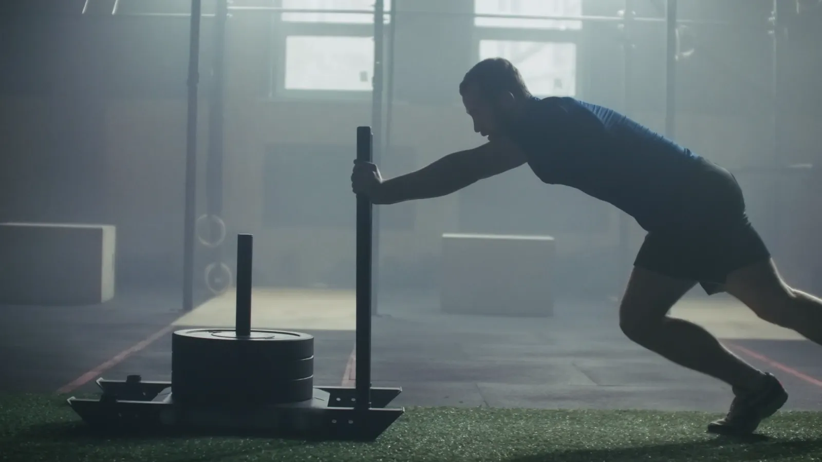 An athlete driving a loaded sled across the turf in a dim gym, the Hyrox sled push