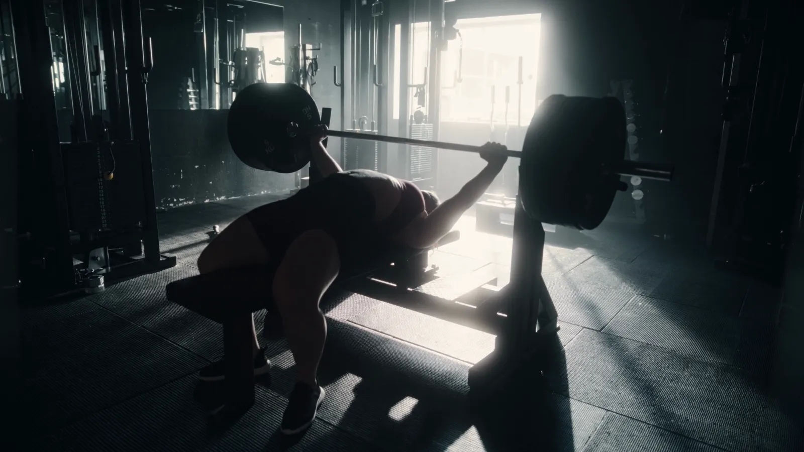 A lifter pressing a loaded barbell on a bench, backlit by gym windows, the heavy press on push day